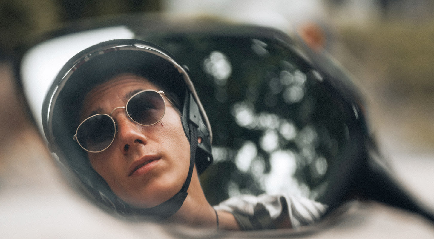 Close-up of a person with glasses and a helmet, reflected in a motorcycle mirror. Trees and a blurred background are visible.